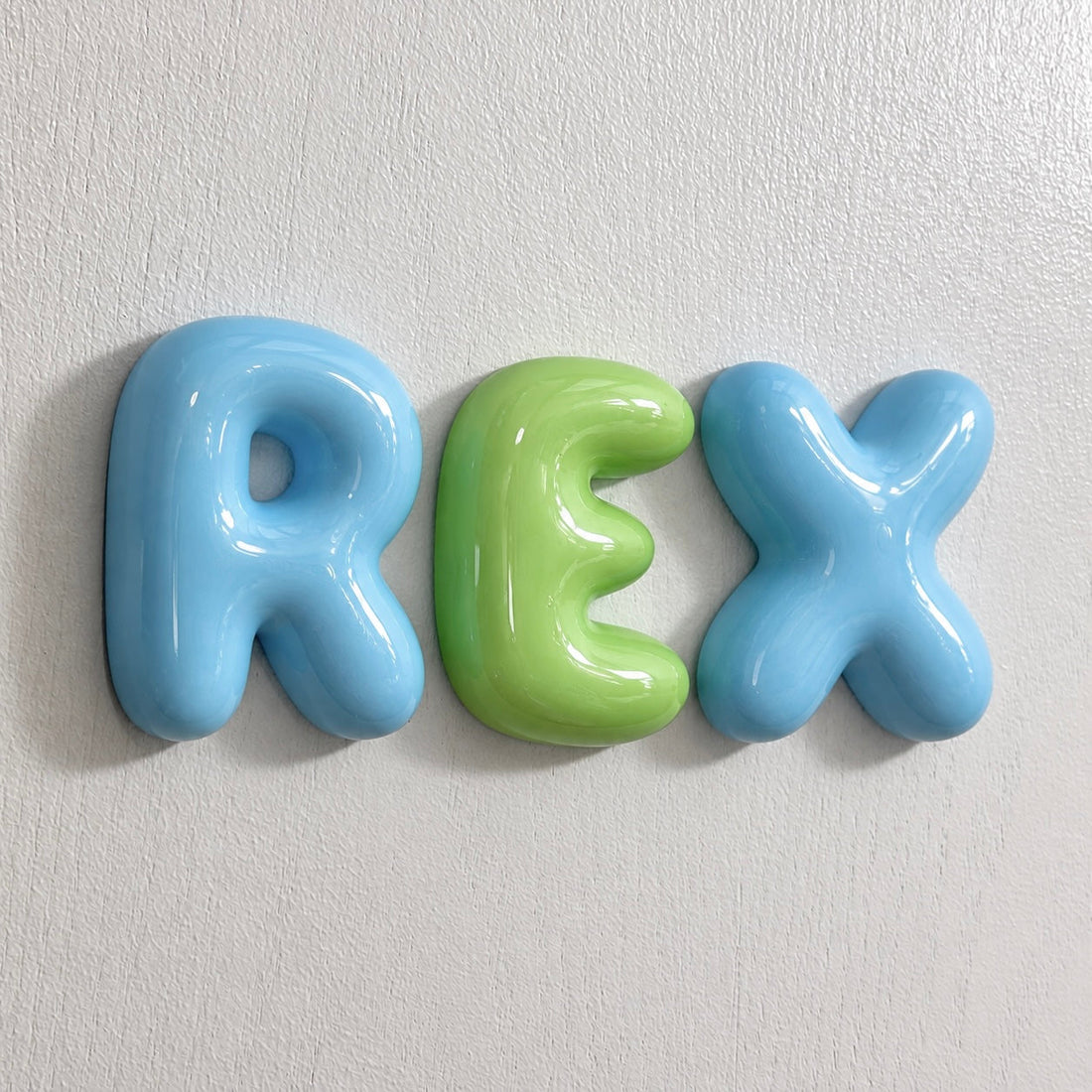 Glossy blue and green 3D bubble letters spelling REX mounted on a wall.