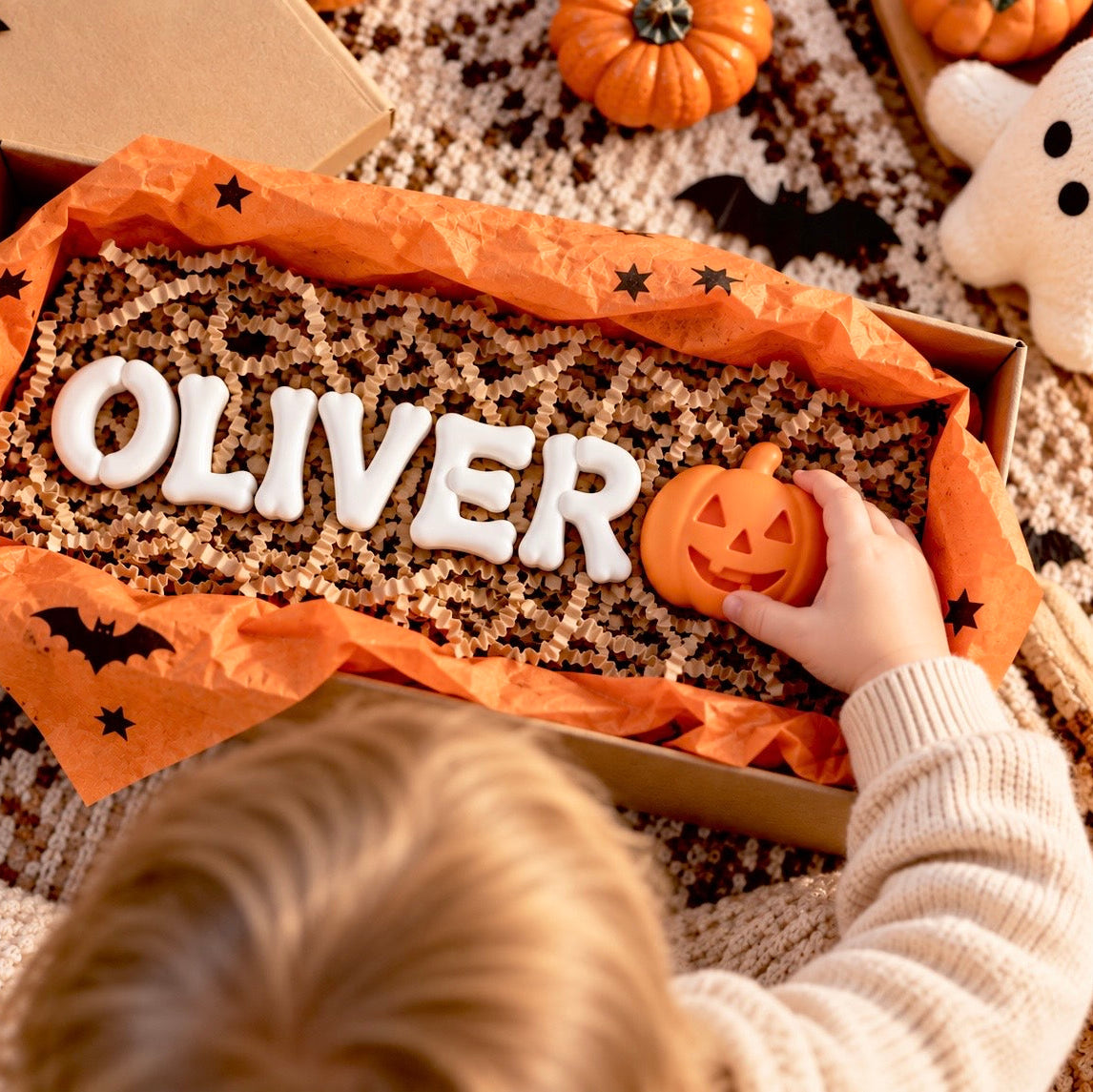 Personalized Halloween gift box with white 3D bone name letters spelling OLIVER and an orange pumpkin accent, shown with a child’s hand.