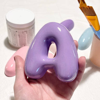 Hand holding a glossy purple 3D bubble letter A with a ceramic-look finish