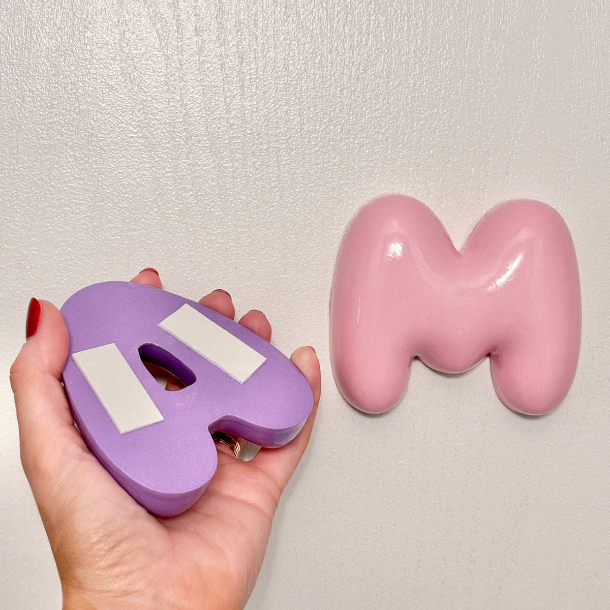 Back view of a purple 3D bubble letter with pre-applied adhesive strips, next to a glossy pink personalized nursery wall letter mounted on the wall.