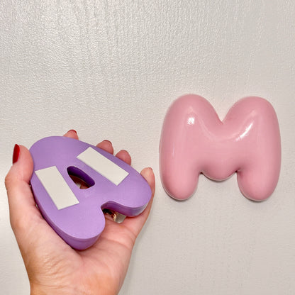 Back view of a purple 3D bubble letter with pre-applied adhesive strips, next to a glossy pink personalized nursery wall letter mounted on the wall.