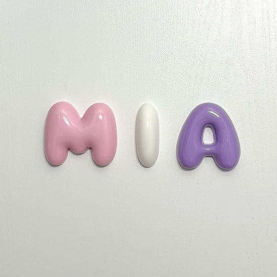 Close-up video of personalized glossy bubble letters spelling MIA in pink, white and purple
