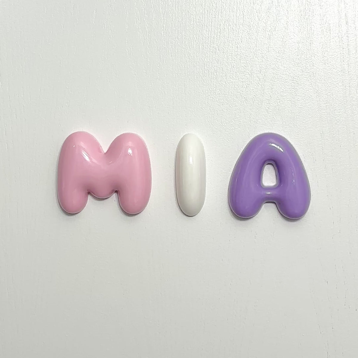 Close-up video of personalized glossy bubble letters spelling MIA in pink, white and purple
